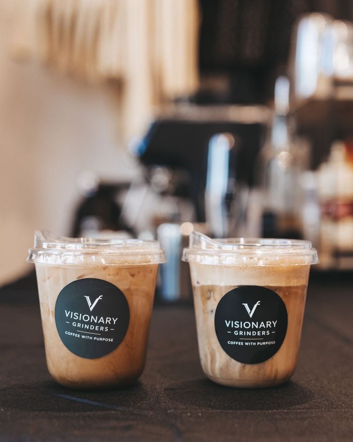 Two iced lattes with Visionary Grinders labels on a bar